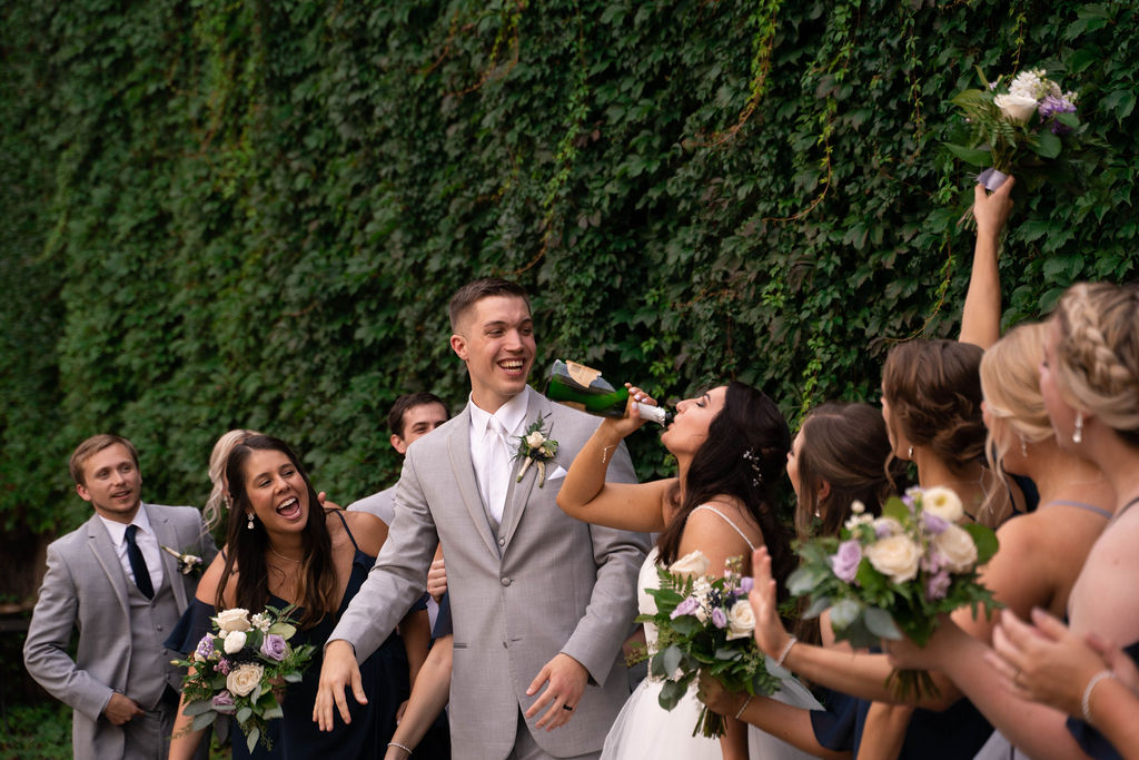 Wedding Party and groom cheer on bride as she chugs champagne straight from the bottle